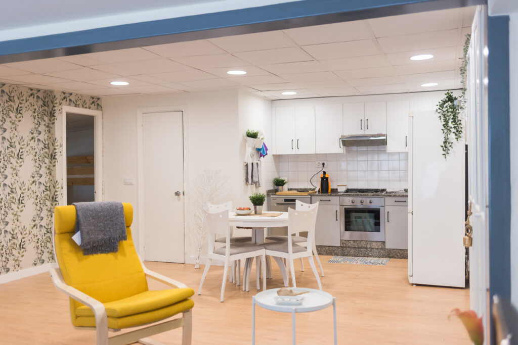 Cocina y comedor abiertos con mesa blanca, sillón amarillo y electrodomésticos